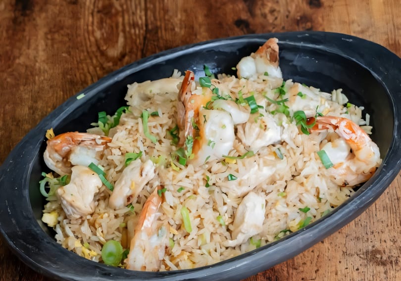 Shrimp and chicken fried rice with chopped green onions in a black bowl on a wooden table.