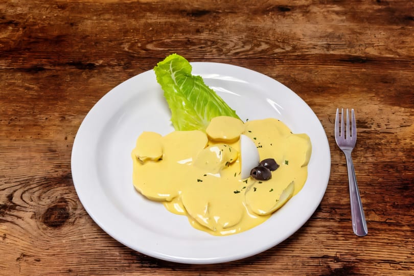 Potatoes covered in yellow huancaína sauce, garnished with lettuce, egg, and olives on a white plate.